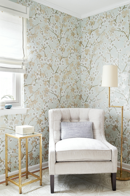 Wallpaper with aqua background and gold and white cherry blossoms behind a cream chair with a brass and glass side table and a brass floor lamp