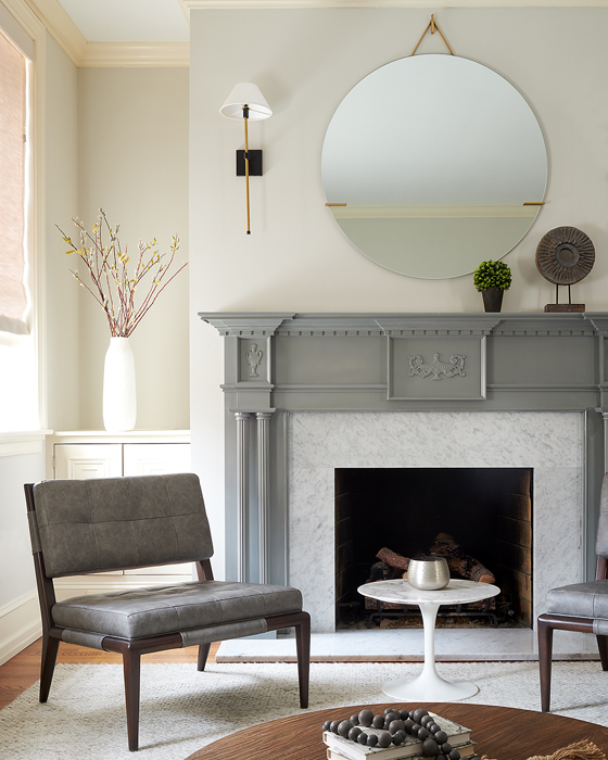 A fireplace with a gray painted wooden mantle featuring carved details and a marble slab hearth and surround, with a round mirror above 