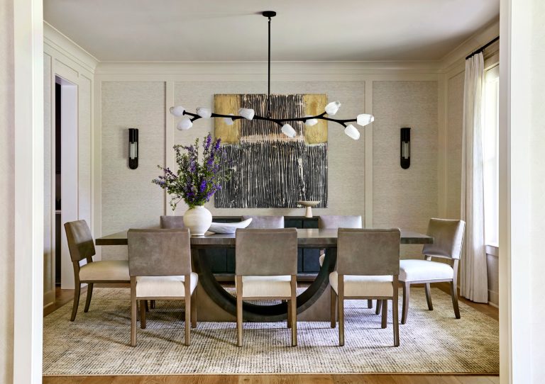 Modern dining room located in Bucks County with a round gray table, six beige upholstered chairs, a large vase with purple flowers, and abstract art on the wall. Interior designer Glenna Stone’s touch is seen in every elegant detail.