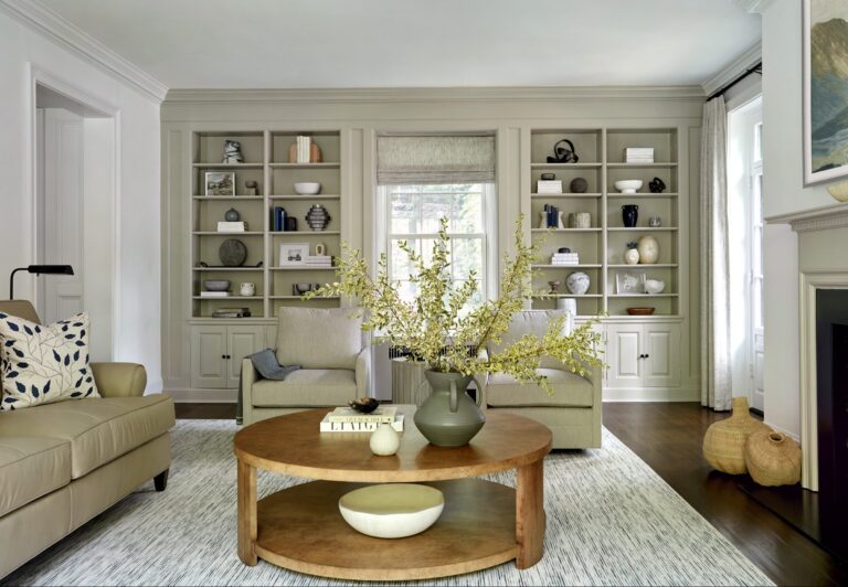 A bright living room designed by interior designer Glenna Stone features beige walls, built-in shelves, and a round wooden coffee table. Located in Chestnut Hill East near Philadelphia, the space is filled with natural light from large windows.