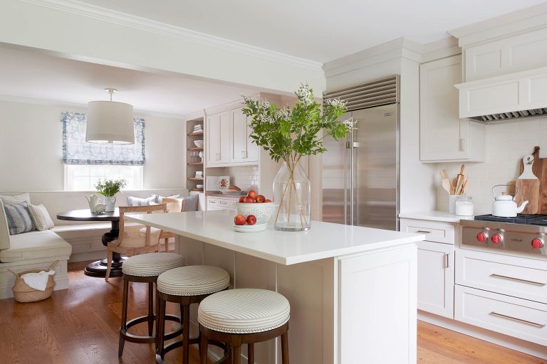 A bright, modern kitchen by interior designer Glenna Stone, located in Lafayette Hill, features white cabinets, a large island with three round stools, stainless steel appliances, hardwood floors, and a cozy breakfast nook in the background.