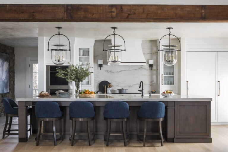 This modern kitchen, located in New Hope in Bucks County, features a large island with four blue stools, pendant lights, a marble backsplash, stainless steel appliances, white cabinetry, and décor curated by interior designer Glenna Stone.