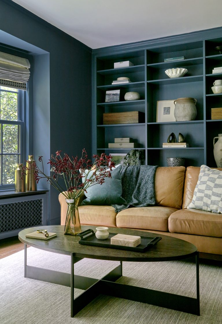 A cozy living room by interior designer Glenna Stone features a tan leather sofa, plaid and knit pillows, red flowers on a modern coffee table, and navy blue bookshelves. Sunlight streams through the window in this Chestnut Hill East home near Philadelphia.