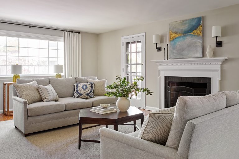 A bright living room designed by interior designer Glenna Stone, located in Lafayette Hill, features two light gray sofas, a dark wood coffee table, a white fireplace with modern art above it, sconces, large windows with white curtains, and a green plant centerpiece.