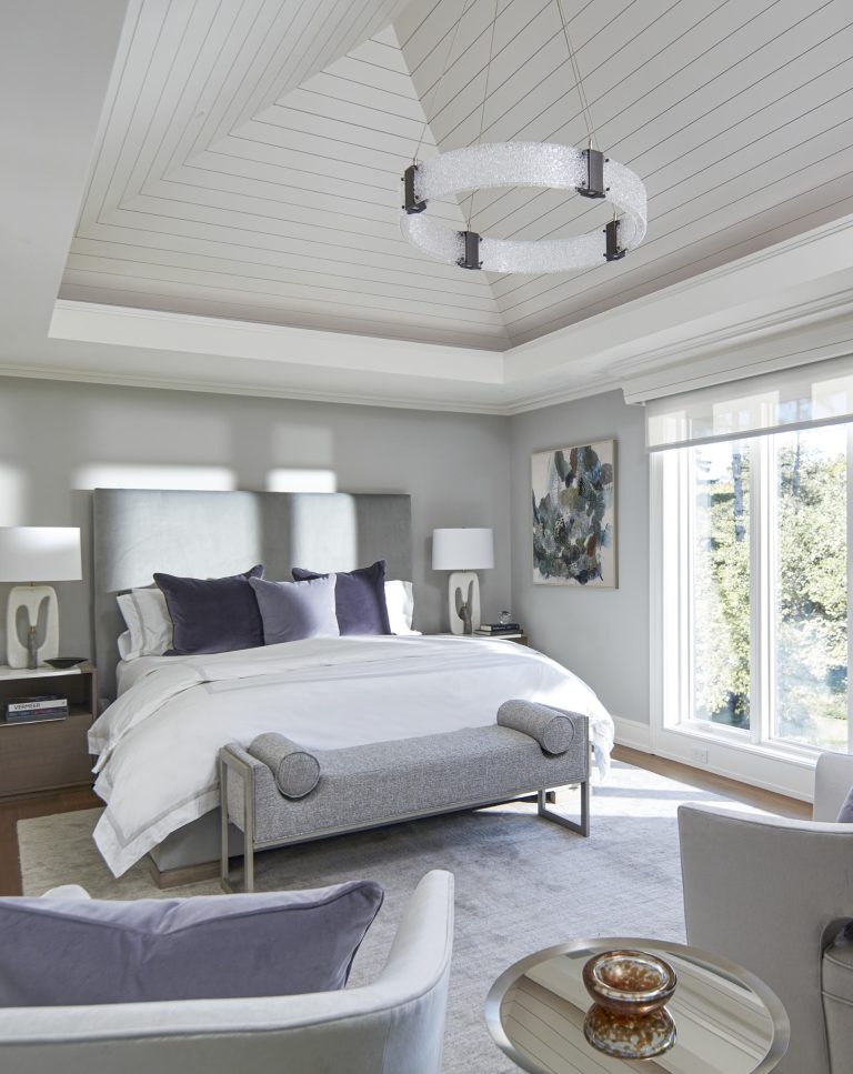 A modern bedroom located on the Main Line features a vaulted, white wood-paneled ceiling, a gray upholstered bed with stylish bedding, cozy armchairs, and large windows—designed with care by interior designer Glenna Stone.