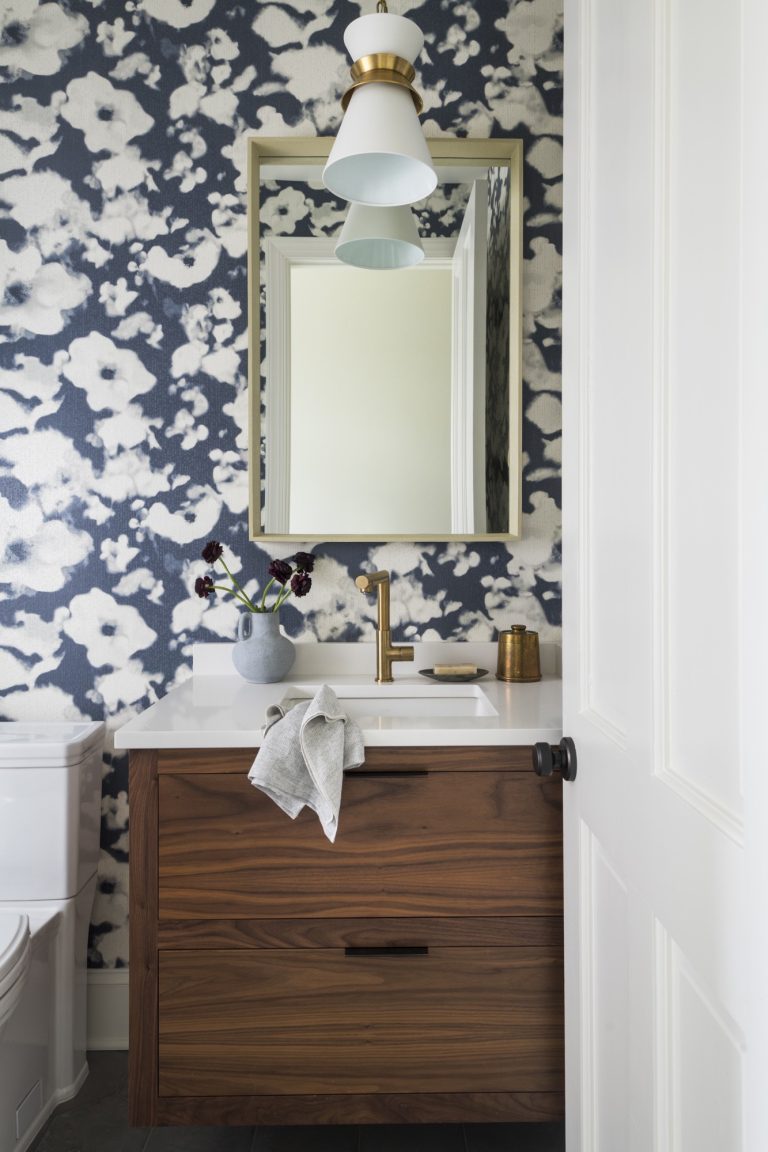 A modern bathroom, designed by interior designer Glenna Stone and located in Gladwyne on the Main Line, features dark floral wallpaper, a wooden vanity with a white countertop, gold accents, a square mirror with gold frame, and elegant touches throughout.