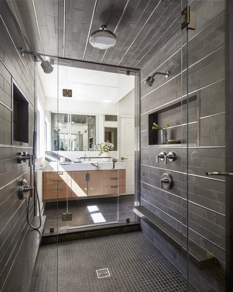 Modern bathroom located on the Main Line, with a glass-enclosed shower featuring gray tile walls and floor, dual shower heads, and stainless steel fixtures. Designed by interior designer Glenna Stone, the space is bright with natural light and elegant finishes.