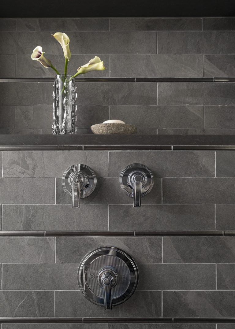A modern shower wall with gray rectangular tiles, designed by interior designer Glenna Stone and located on the Main Line, features three chrome handles and a clear glass vase with white calla lilies beside a smooth stone for an elegant, minimalist look.