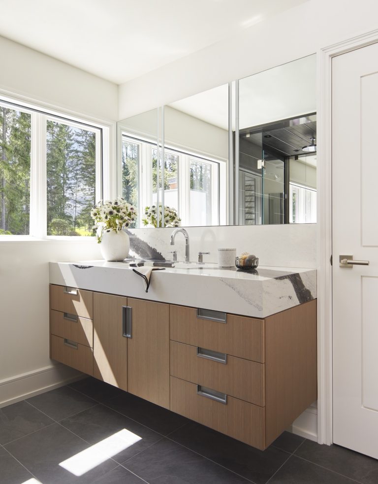 A modern bathroom designed by interior designer Glenna Stone features a floating wooden vanity, a marbled countertop, and sleek accents. Large mirrors and windows flood the Main Line space with natural light, highlighting its clean, minimalist style.