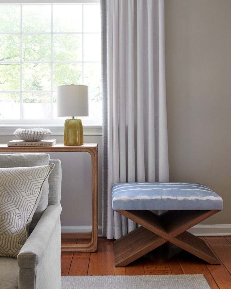Bright room located in Lafayette Hill featuring a large window, white curtains, and wooden floor. Interior designer Glenna Stone styled a small wooden table with a yellow lamp and bowl beside a cozy gray sofa with geometric pillows.