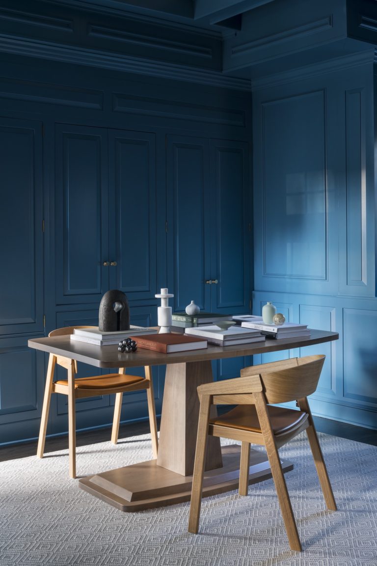 A modern room by interior designer Glenna Stone features blue paneled walls, a light wood table with two chairs, and stacked books. Soft light casts gentle shadows across the textured off-white rug in this Main Line Gladwyne home.