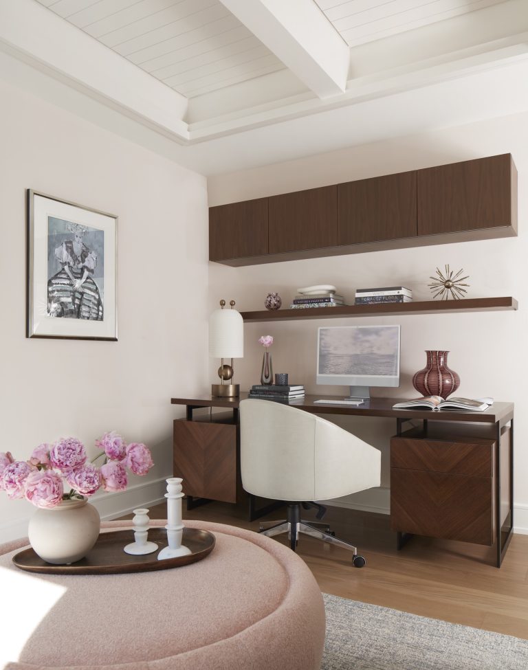 A modern home office located on the Main Line features a dark wood desk, white swivel chair, and sleek accessories. Shelves with decor line the wall, while pink flowers in a vase add a touch of color—styled by interior designer Glenna Stone.