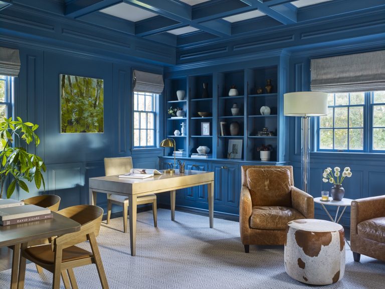 Sophisticated home office, designed by interior designer Glenna Stone, with blue paneled walls, built-in shelves, leather armchairs, cream desk, potted plant, modern art, and large windows with Roman shades. Neutral rug covers the floor.