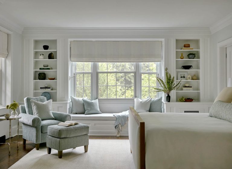 A serene bedroom designed by interior designer Glenna Stone features large windows framed by white built-ins, a cushioned window seat, a blue armchair, and soft daylight—located in Chestnut Hill East near Philadelphia.