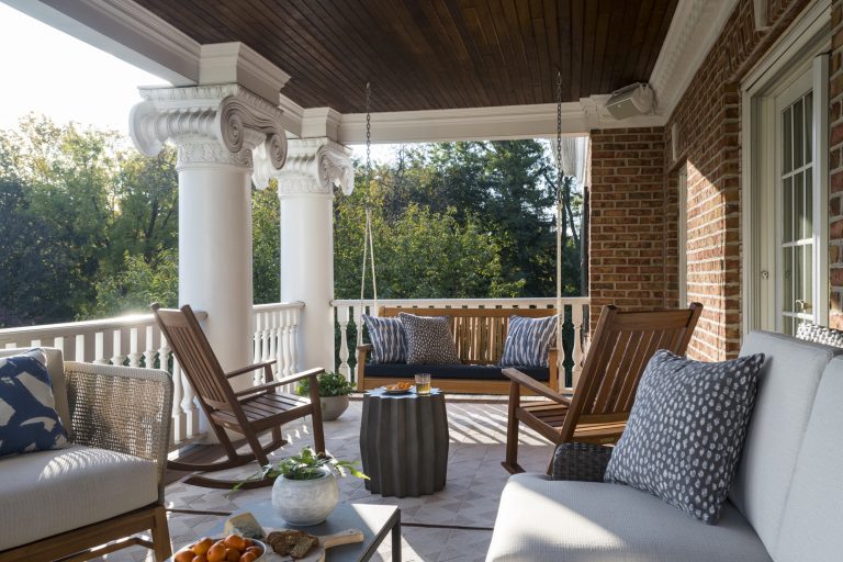 A stylish covered porch located in Gladwyne on the Main Line, with white columns, ornate capitals, a wooden ceiling, and brick walls. It features a porch swing, wooden chairs, a cushioned sofa with patterned pillows, and views of green trees.