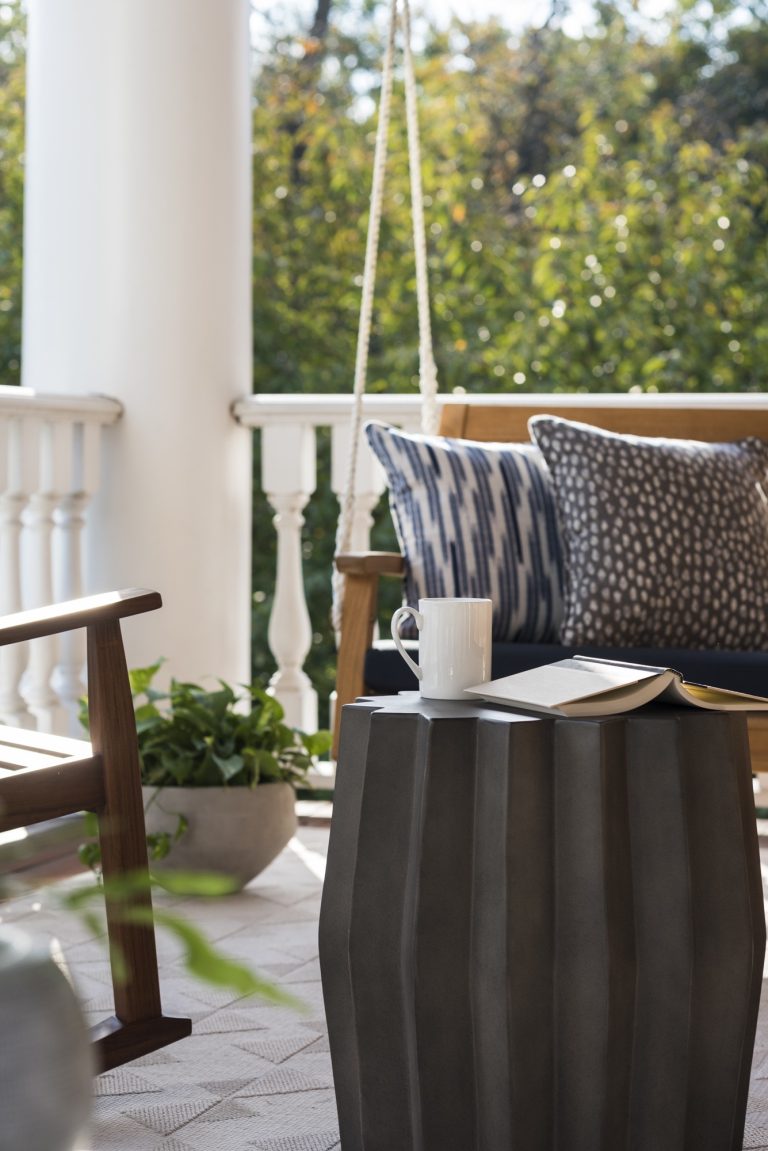 A cozy porch scene located in Gladwyne on the Main Line: a wooden swing with blue and white pillows, fluted metal side table holding a mug and book, rocking chair, potted plant, white railing, and trees in the background.
