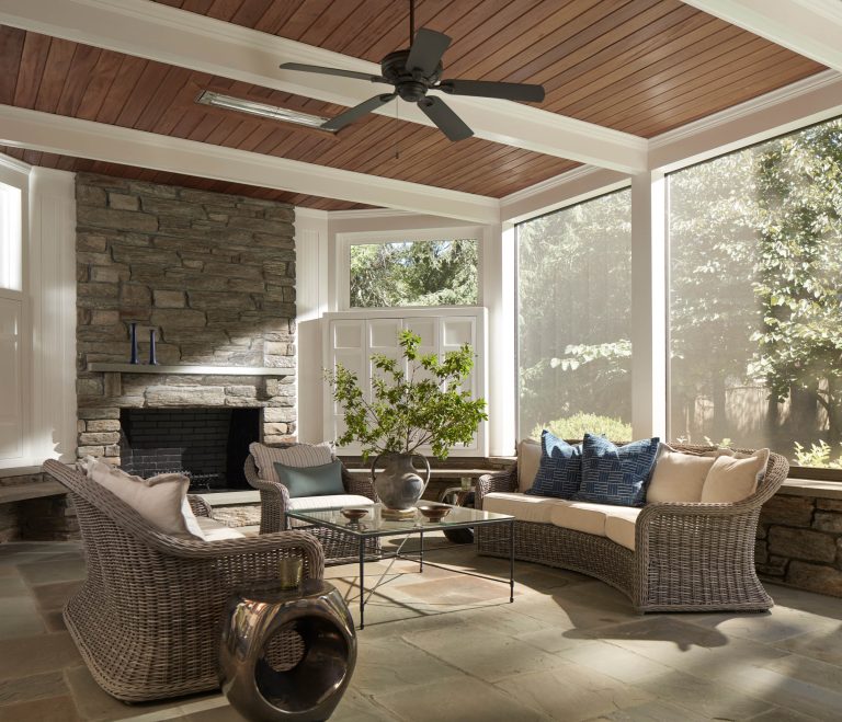 Located on the Main Line, this sunroom with wicker sofas and blue and cream cushions by interior designer Glenna Stone features a stone fireplace, screened windows, and wooden ceiling, creating a serene, cozy atmosphere filled with sunlight.