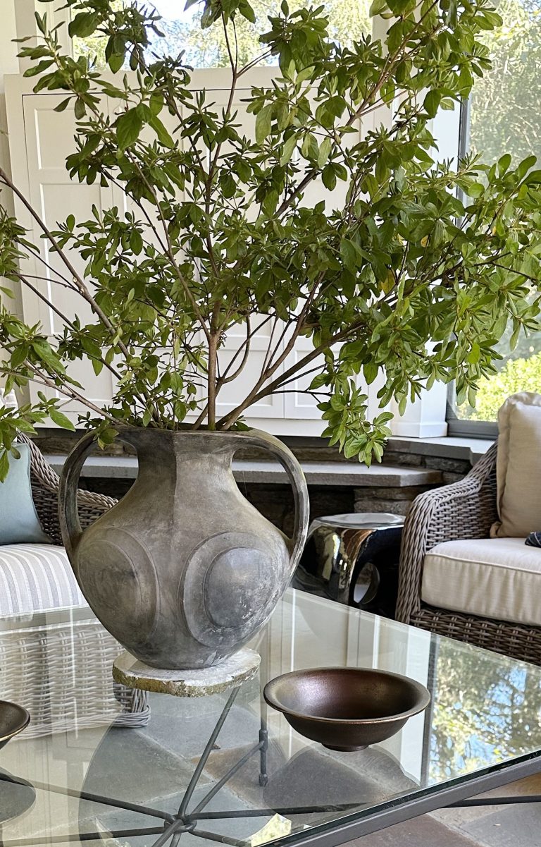 A large, round, grayish vase with handles holds leafy green branches on a glass table. The airy living space, designed by interior designer Glenna Stone, features wicker armchairs and is filled with sunlight from large windows.