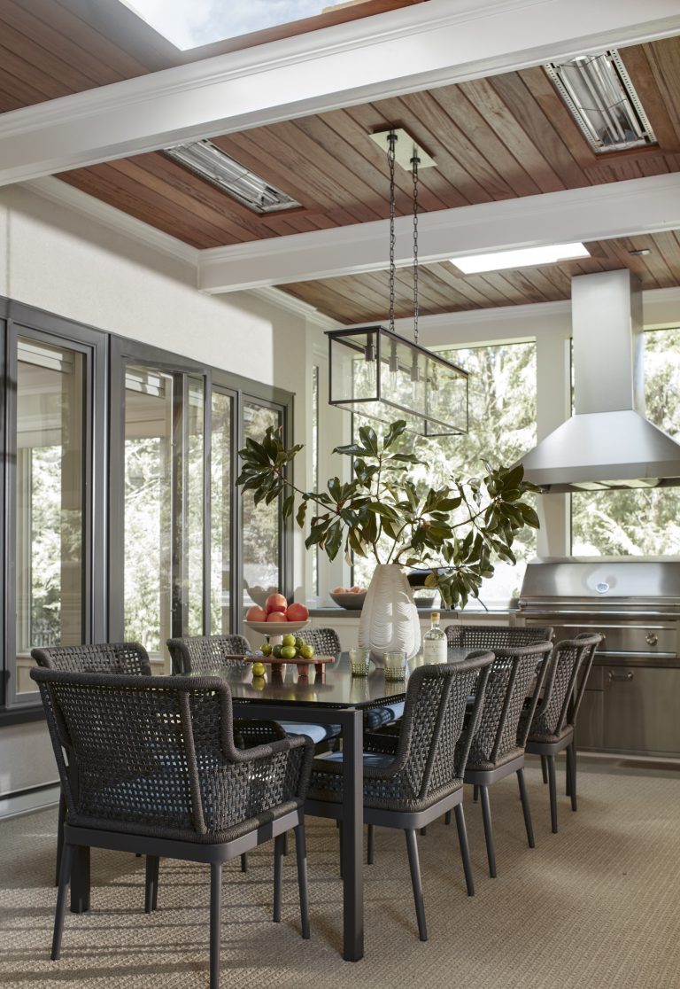 A modern sunroom dining area, designed by interior designer Glenna Stone, features a large black table, woven chairs, and a stainless steel grill. Expansive windows showcase greenery in this Main Line home with wood-paneled ceiling and sleek light fixtures.