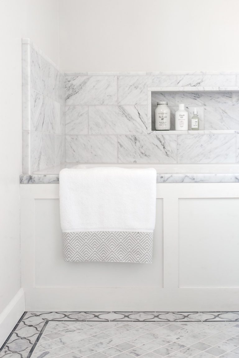 A white and gray bathroom, located in Lafayette Hill, features a marble-tiled bathtub with a white towel draped over the side. The space, styled by interior designer Glenna Stone, includes built-in shelves and matching gray marble with geometric patterns.