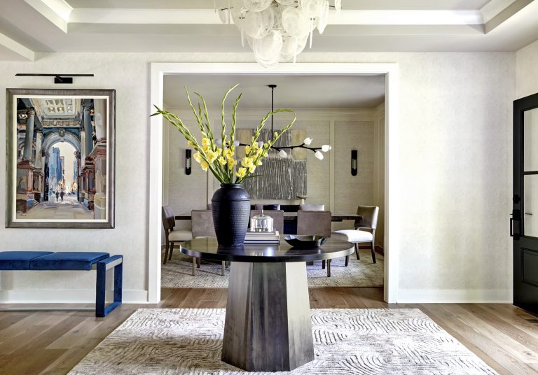 A modern entryway by interior designer Glenna Stone features a round table with a black vase and tall yellow flowers on a textured rug. A dining area, wall art, blue bench, and sculptural chandelier complete this Bucks County home.