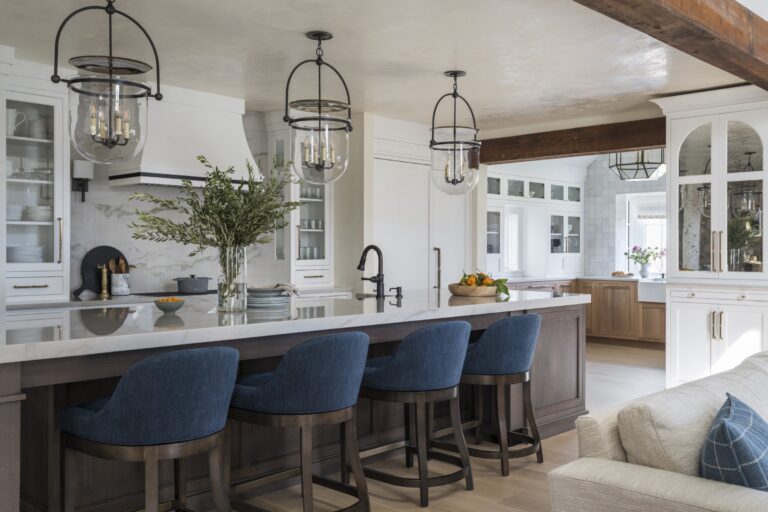 A modern kitchen located in New Hope in Bucks County features a large island with four blue upholstered stools, a white countertop, and a black faucet. Pendant lights, white cabinetry, glass-front cabinets, and greenery in a vase complete the space.
