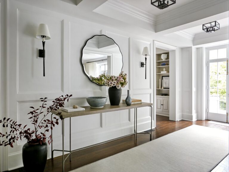 A bright entryway designed by interior designer Glenna Stone features white paneled walls, a wavy-edged round mirror, and a slim console table with vases and decor. Located in Chestnut Hill East near Philadelphia, it boasts wooden floors and wall sconces.
