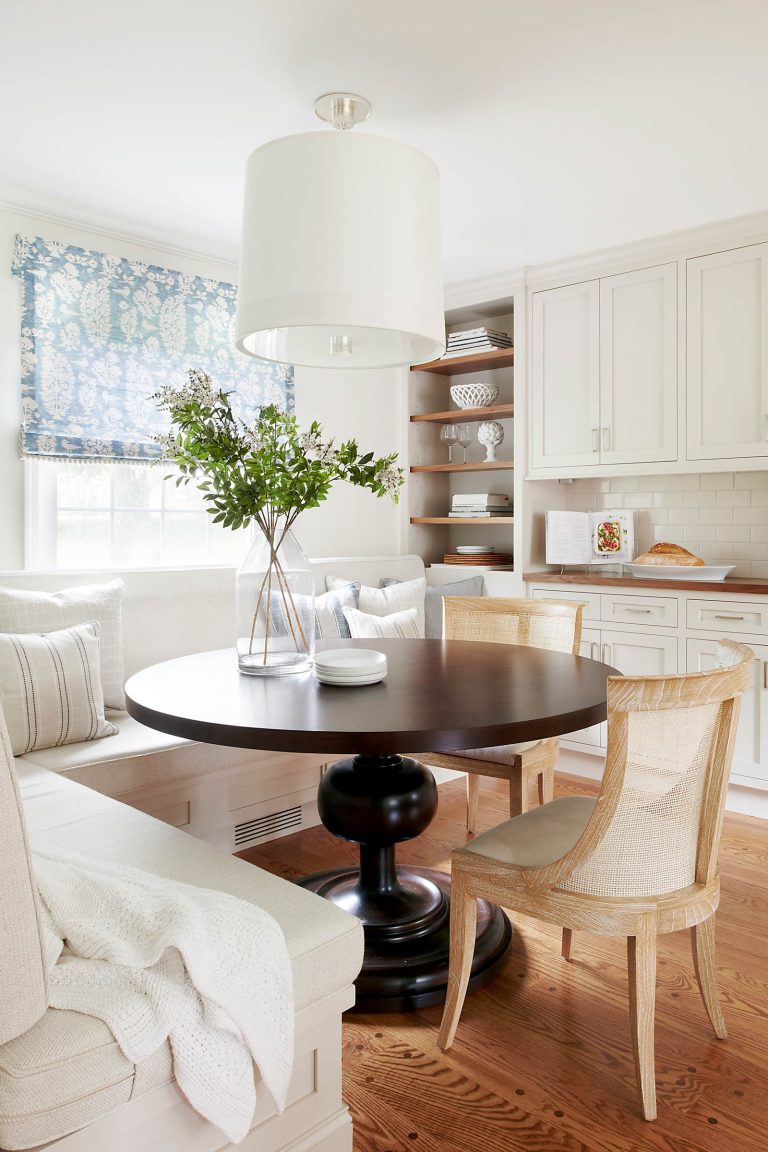 A cozy breakfast nook designed by interior designer Glenna Stone features a round dark wood table, two light wicker chairs, and a built-in bench with white cushions. White cabinets, open shelves, and a large drum pendant complete this Lafayette Hill space.
