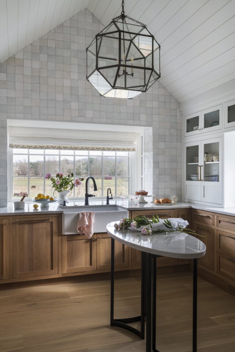 A bright, modern farmhouse kitchen located in New Hope in Bucks County with wood cabinets, white countertops, and a farmhouse sink under a large window. A geometric pendant light hangs above a marble island adorned with flowers and a knife.