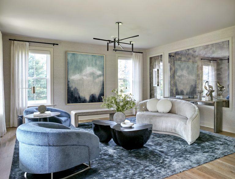 Modern living room by interior designer Glenna Stone with a large abstract painting, blue velvet chairs, a curved beige sofa, sculptural coffee tables, and a textured blue rug. Light streams through tall windows in this Bucks County retreat.