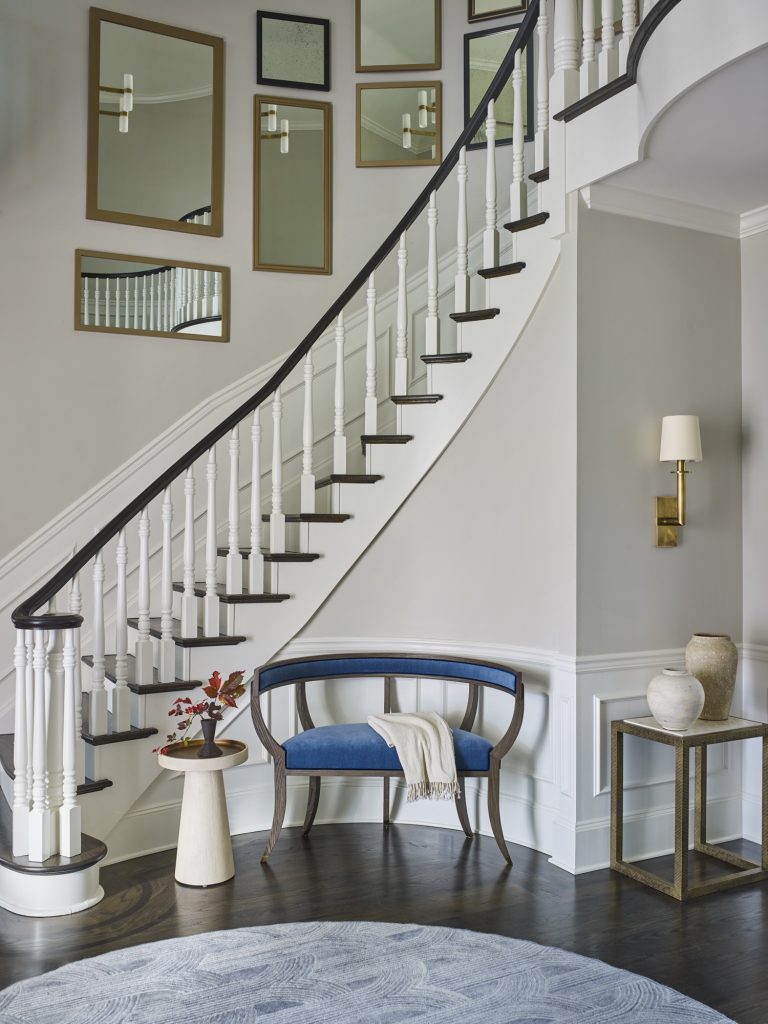 A curved staircase with white spindles and dark wood railing winds upward in this Gladwyne home on the Main Line, designed by interior designer Glenna Stone. A blue bench, mirrors, and a side table with red flowers add modern touches and brighten the space.