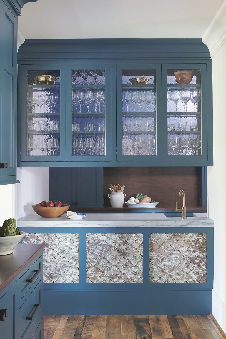 A blue kitchen designed by interior designer Glenna Stone features glass-front upper cabinets holding glassware, a marble countertop, gold faucet, and a lower cabinet with textured glass. Natural wood floors anchor this elegant space located in New Hope in Bucks County.