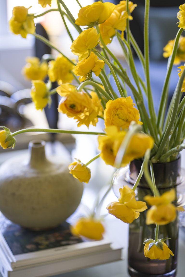 A vase holds a bouquet of bright yellow flowers with long green stems, arranged loosely on a table. Next to it is a round ceramic vase and a stack of books, reflecting the elegant style of interior designer Glenna Stone, located on the Main Line.