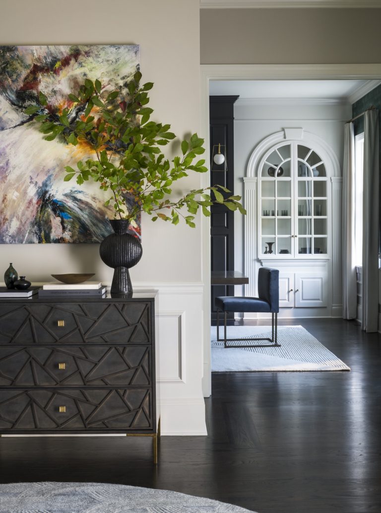 Modern interior by Glenna Stone, located in Gladwyne on the Main Line, features a dark geometric dresser, black vase with green branches, abstract painting above, and a bright room with arched shelves, window, table, and blue chair on a light rug.