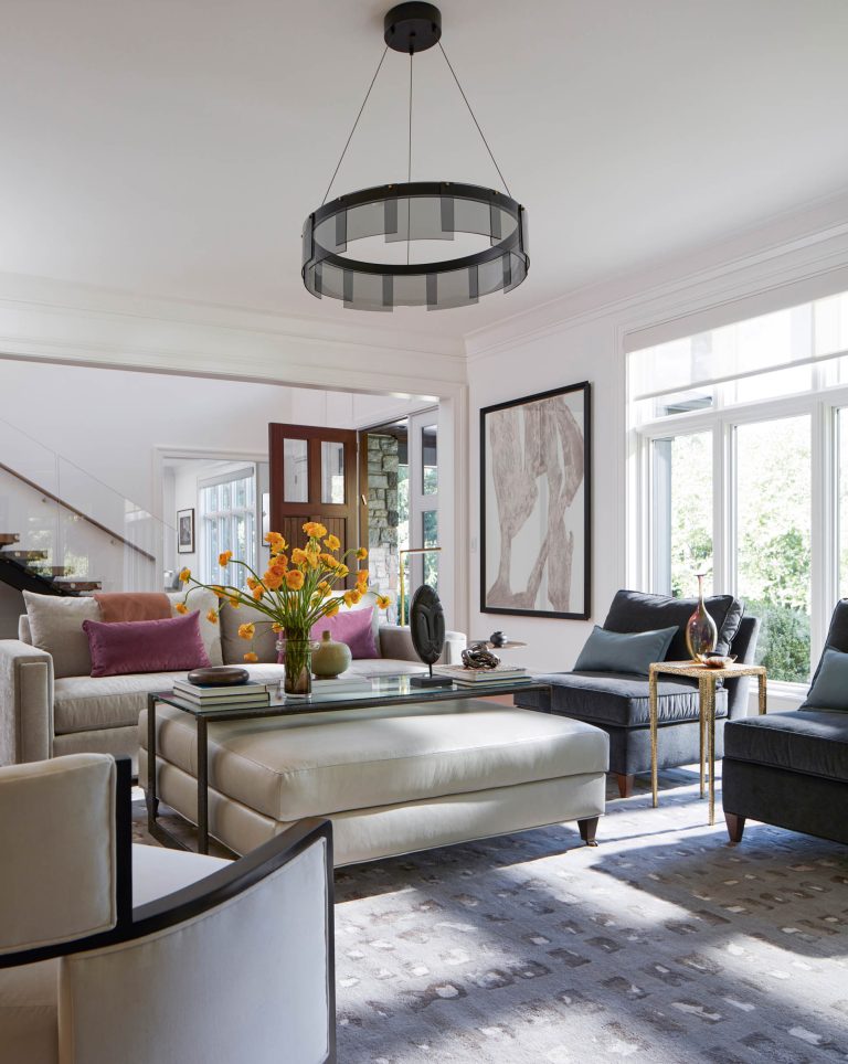 Modern living room by interior designer Glenna Stone, located on the Main Line, featuring large windows, neutral-toned sofas, a glass coffee table with yellow flowers, accent chairs, plush cushions, contemporary art, and a round black chandelier. Light fills the space.