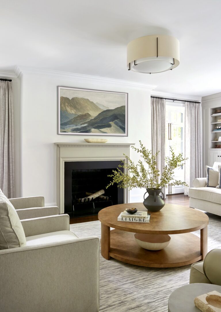 A modern living room by interior designer Glenna Stone, located in Chestnut Hill East near Philadelphia, features light gray armchairs, a round wooden coffee table with green branches, a beige rug, a fireplace with art above, and built-in shelves.