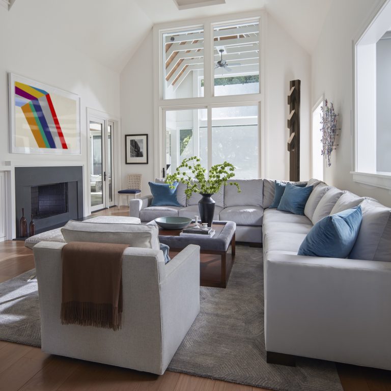 A modern living room by interior designer Glenna Stone, located on the Main Line, features a large gray sectional with blue accent pillows, a cozy armchair, stylish coffee table with plants, gray rug, fireplace with colorful abstract art, and sunlit windows.