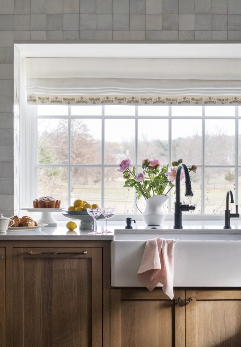 A bright kitchen designed by interior designer Glenna Stone features a large farmhouse sink, black faucet, and wooden cabinets. A big window overlooks a scenic, tree-filled yard, making this New Hope, Bucks County home feel warm and inviting.