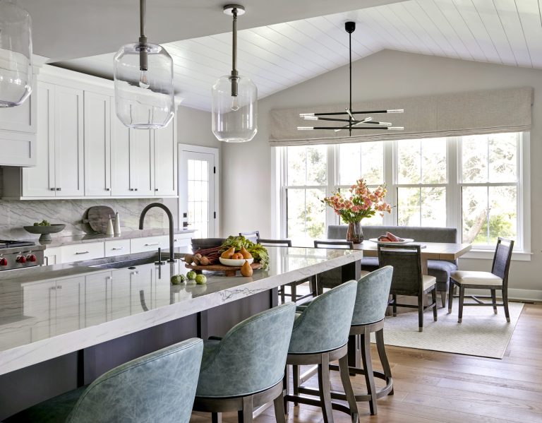 A modern, sunlit kitchen located in Bucks County features a marble island with teal bar stools, pendant lights, and a fruit bowl. White cabinets, a sink, and stove line the back wall. A dining area with flowers overlooks greenery.