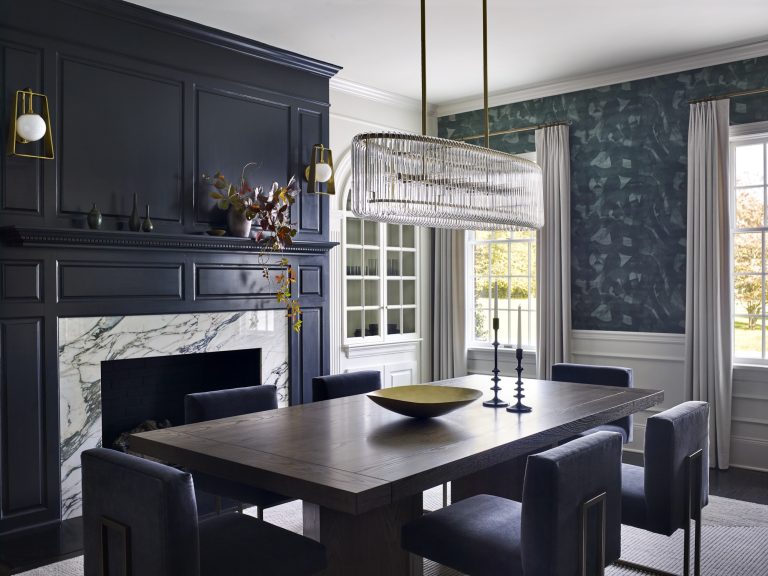 Modern dining room located in Gladwyne on the Main Line, featuring a dark wood table with six cushioned chairs, marble fireplace with black mantle, moody green walls, large windows with white trim, and minimalist décor by interior designer Glenna Stone.