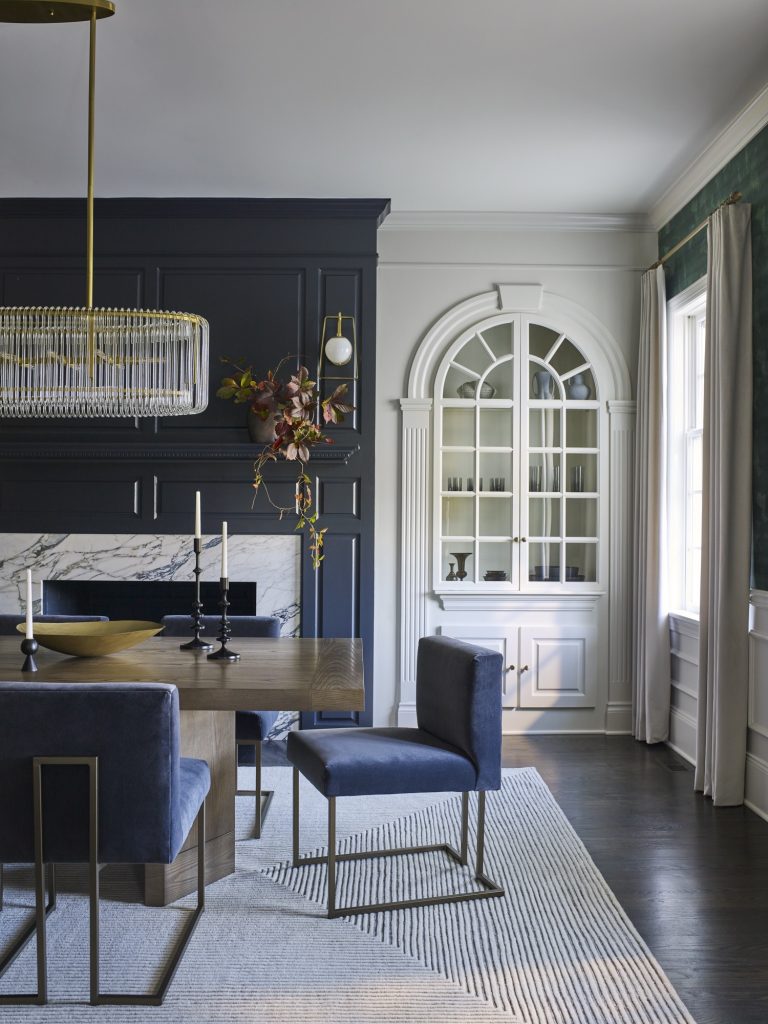 Elegant dining room by interior designer Glenna Stone, located in Gladwyne on the Main Line, features a dark navy accent wall, marble fireplace, modern chandelier, wooden table with blue velvet chairs, arched built-ins, and large bright windows.