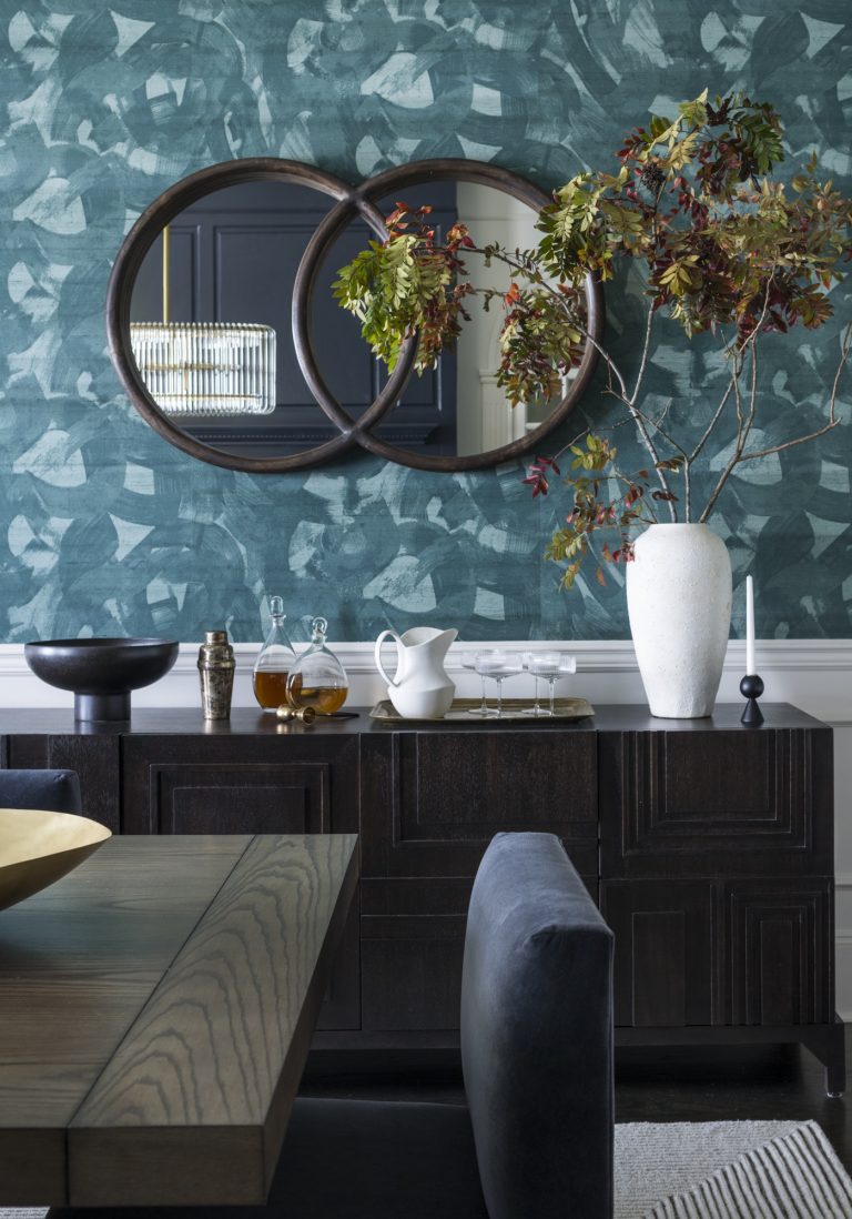 A modern dining room by interior designer Glenna Stone, located in Gladwyne on the Main Line, features a dark wood table and chairs, a stylish sideboard, leafy branches in a white vase, round mirrors, and a striking blue abstract-patterned wall.