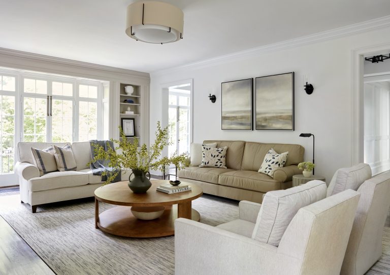 A bright living room designed by interior designer Glenna Stone features large windows, neutral-toned sofas, a round wooden coffee table with greenery, abstract wall art, and soft lighting for a cozy, modern atmosphere.