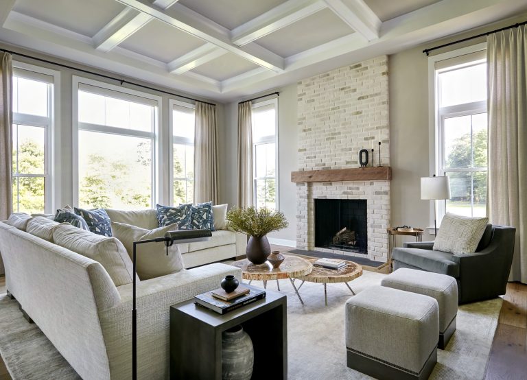 Bright, modern living room located in Bucks County with large windows, light walls, and coffered ceiling. Interior designer Glenna Stone blends neutral sofas and a white brick fireplace with warm decorative accents for an inviting space.
