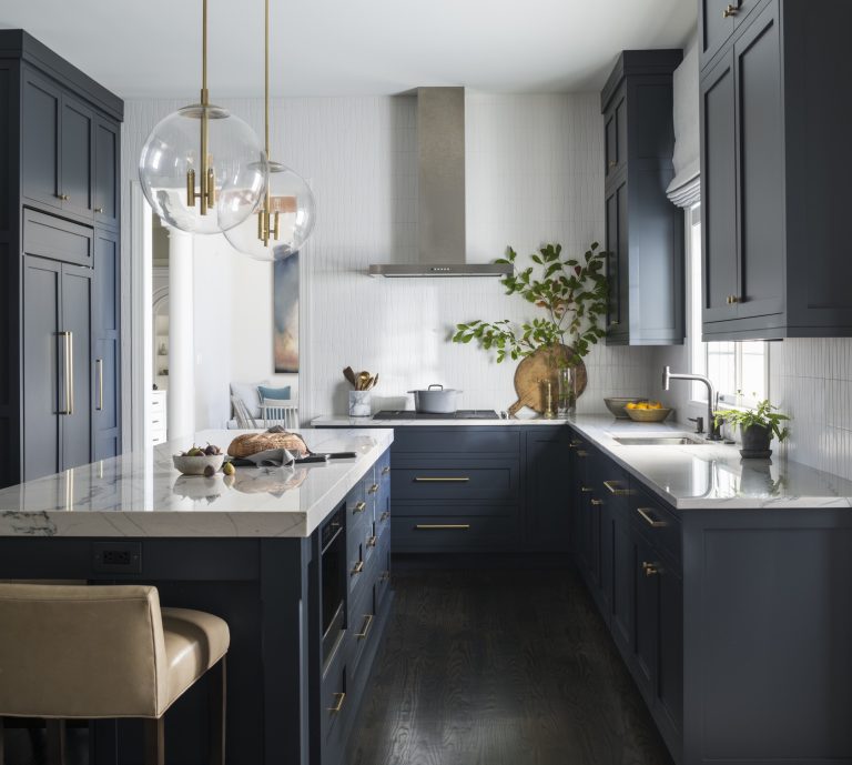 Modern kitchen located in Gladwyne on the Main Line, with dark blue cabinets, brass handles, white marble countertops, and a center island. Designed by interior designer Glenna Stone, it features glass-globe pendants and stainless steel appliances for a warm contrast.
