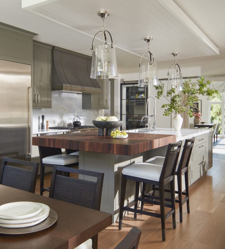 A modern kitchen by interior designer Glenna Stone, located on the Main Line, features gray cabinets, stainless steel appliances, a wood-topped island with bar stools, pendant lights, and a dining table set in sunlit elegance.