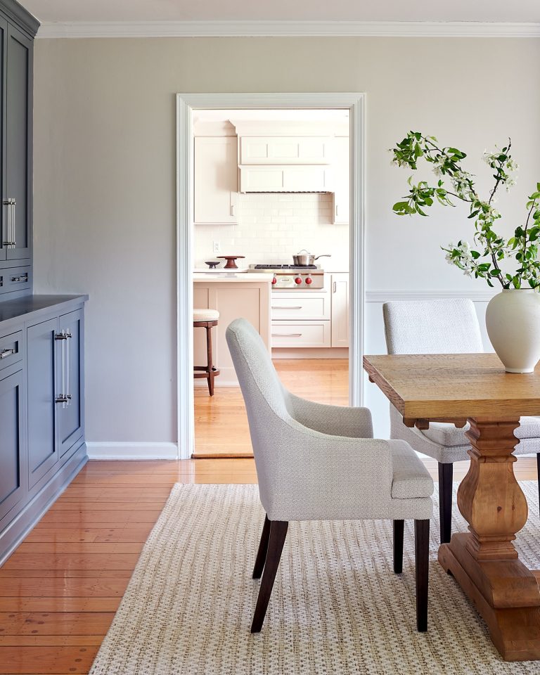 A bright dining room located in Lafayette Hill features a wooden table, upholstered chairs, a vase with green branches, and a woven rug. Designed by interior designer Glenna Stone, the space feels airy and well-lit, with a view into the white kitchen beyond.