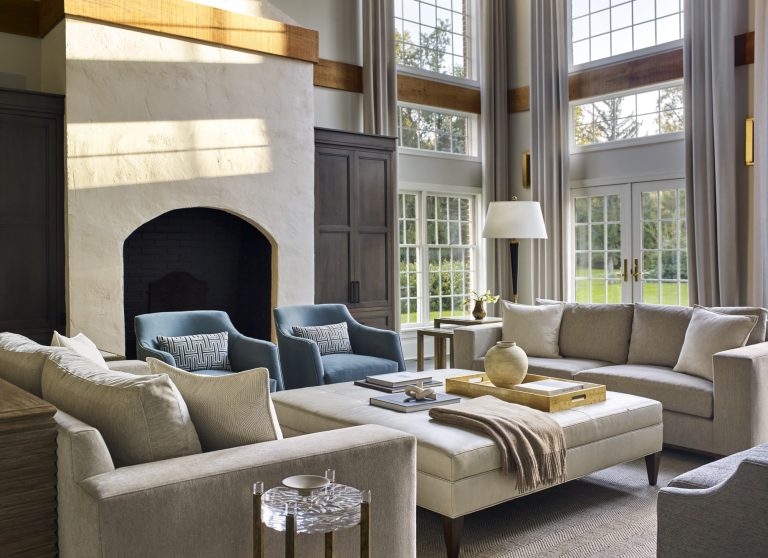 Spacious living room located in Gladwyne on the Main Line, featuring large windows, light gray sectional sofas, blue armchairs, and a white textured fireplace. Designed by interior designer Glenna Stone for an airy, elegant atmosphere with abundant natural light.