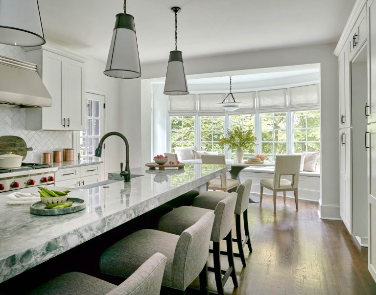 Bright, modern kitchen located in Chestnut Hill East near Philadelphia, featuring a long marble island, gray upholstered stools, pendant lights, and stainless steel appliances. A bay window with cushioned seat invites natural light; fresh flowers and fruit add charm.