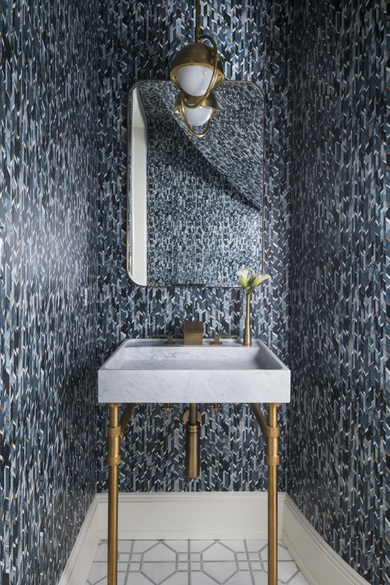 Located in Gladwyne on the Main Line, this small bathroom features geometric dark blue wallpaper, a marble sink with gold fixtures, a gold-framed mirror, and a pendant light. Designed by interior designer Glenna Stone, it’s finished with white geometric floor tiles.
